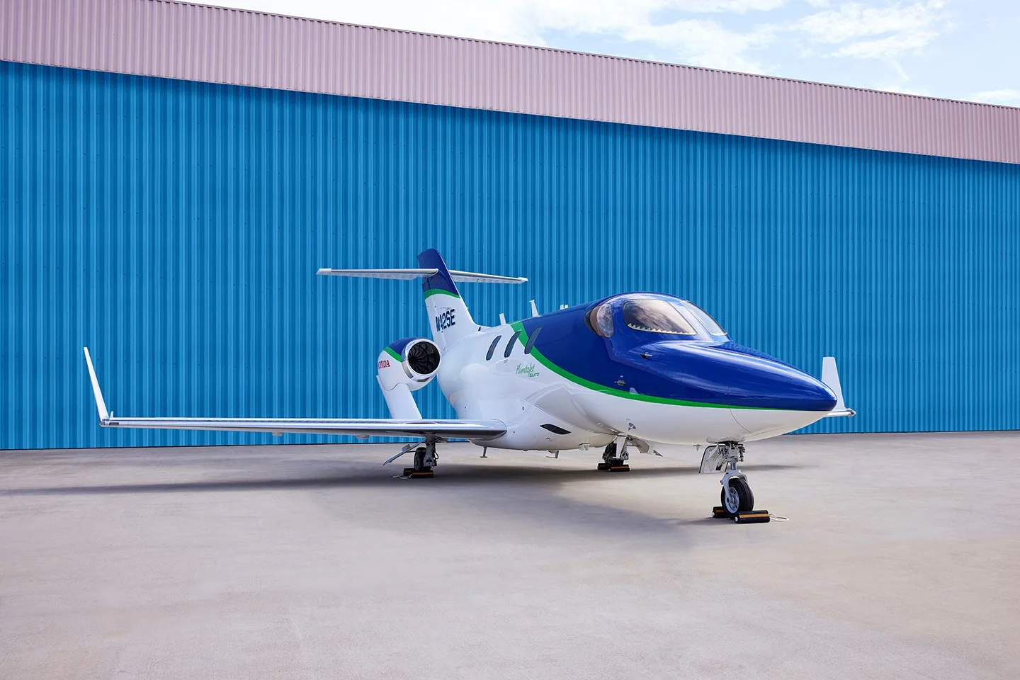A white and blue HondaJet Elite with a green stripe, registration N42SE, parked on an airfield in front of a blue hangar.