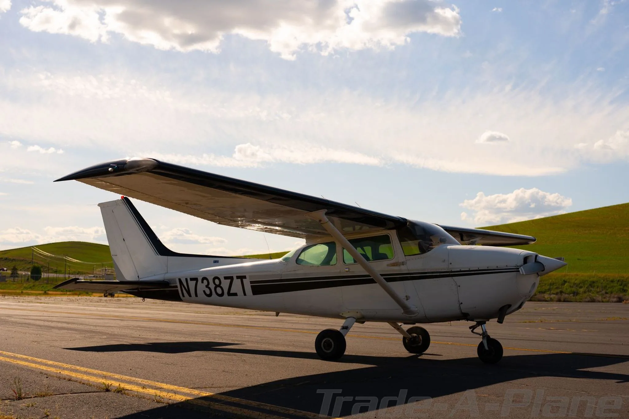 A white Cessna 172 with black stripes, tail number N738ZT, parked on an airfield with green hills in the background.