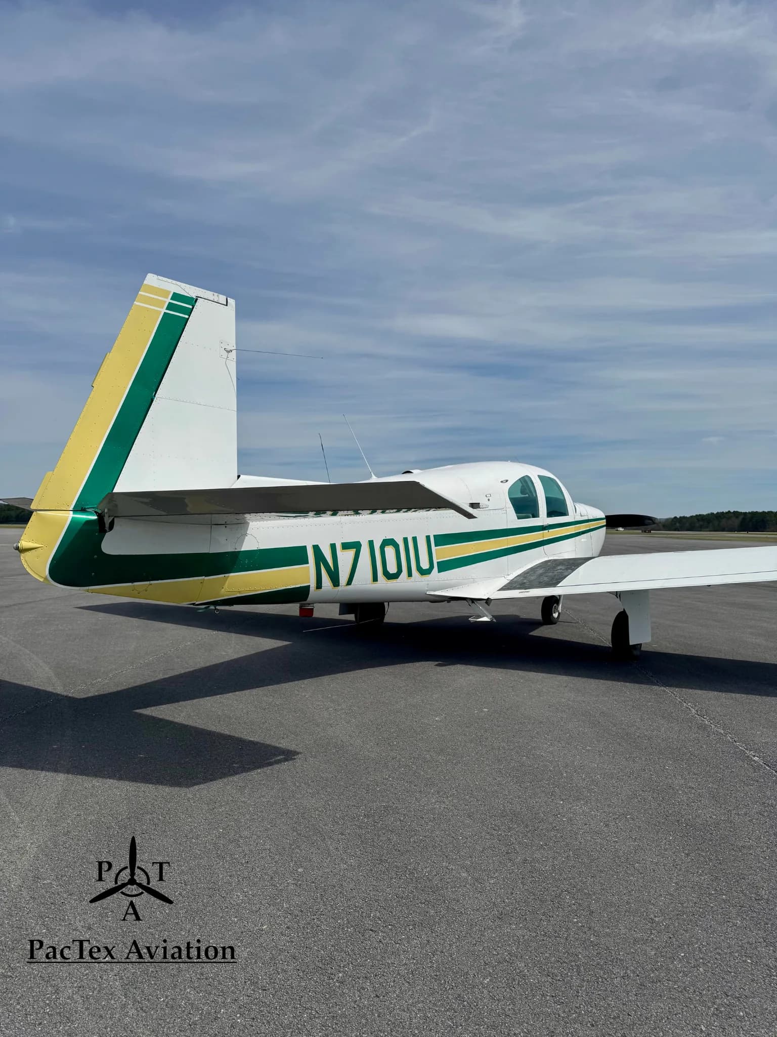 A rear-side view of a white Mooney M20C aircraft with green and yellow stripes, parked on an airport tarmac.