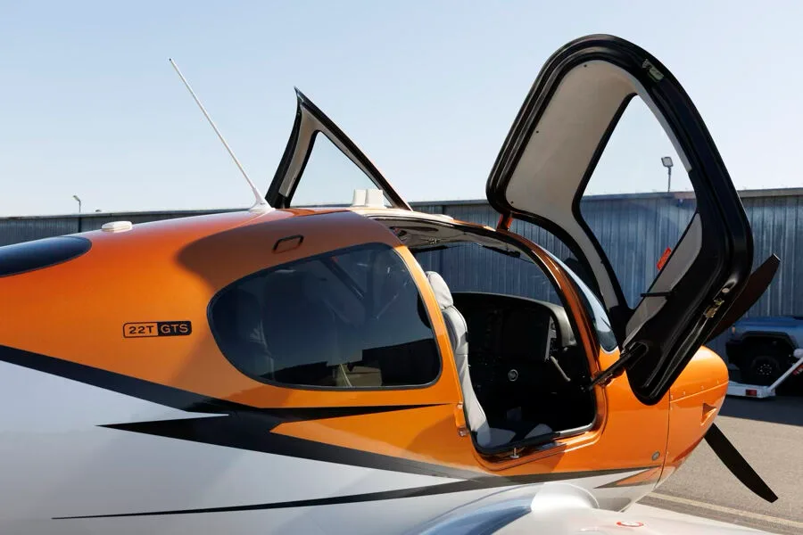 Close-up of the cockpit area of an orange and silver Cirrus SR22T GTS aircraft with both doors open, showing the interior.