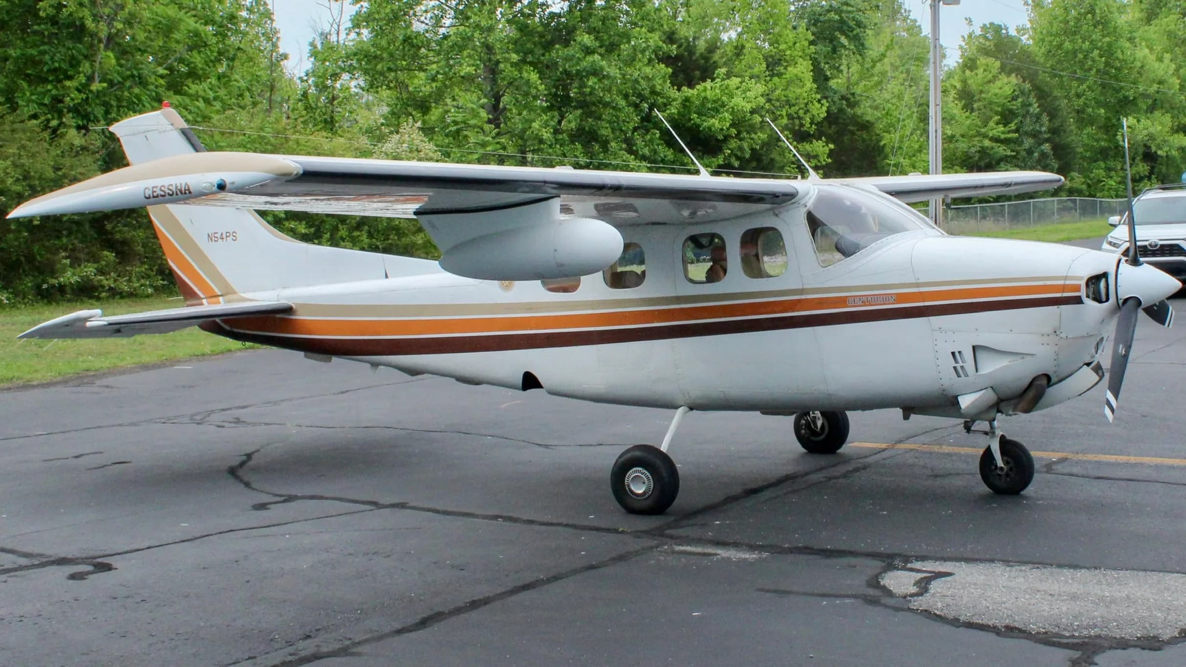 A three-quarter front view of the white Cessna P210N Centurion, registration N54PS, with brown and orange stripes, parked on an apron.