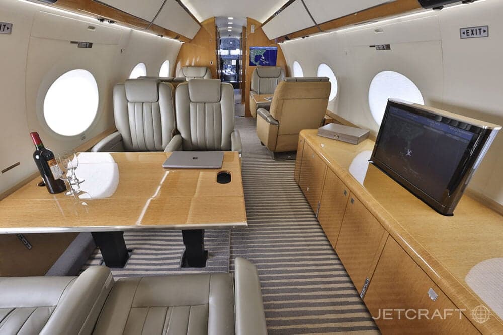 The spacious cabin interior of a business jet with light brown leather seats, a large wooden table, and a television screen.
