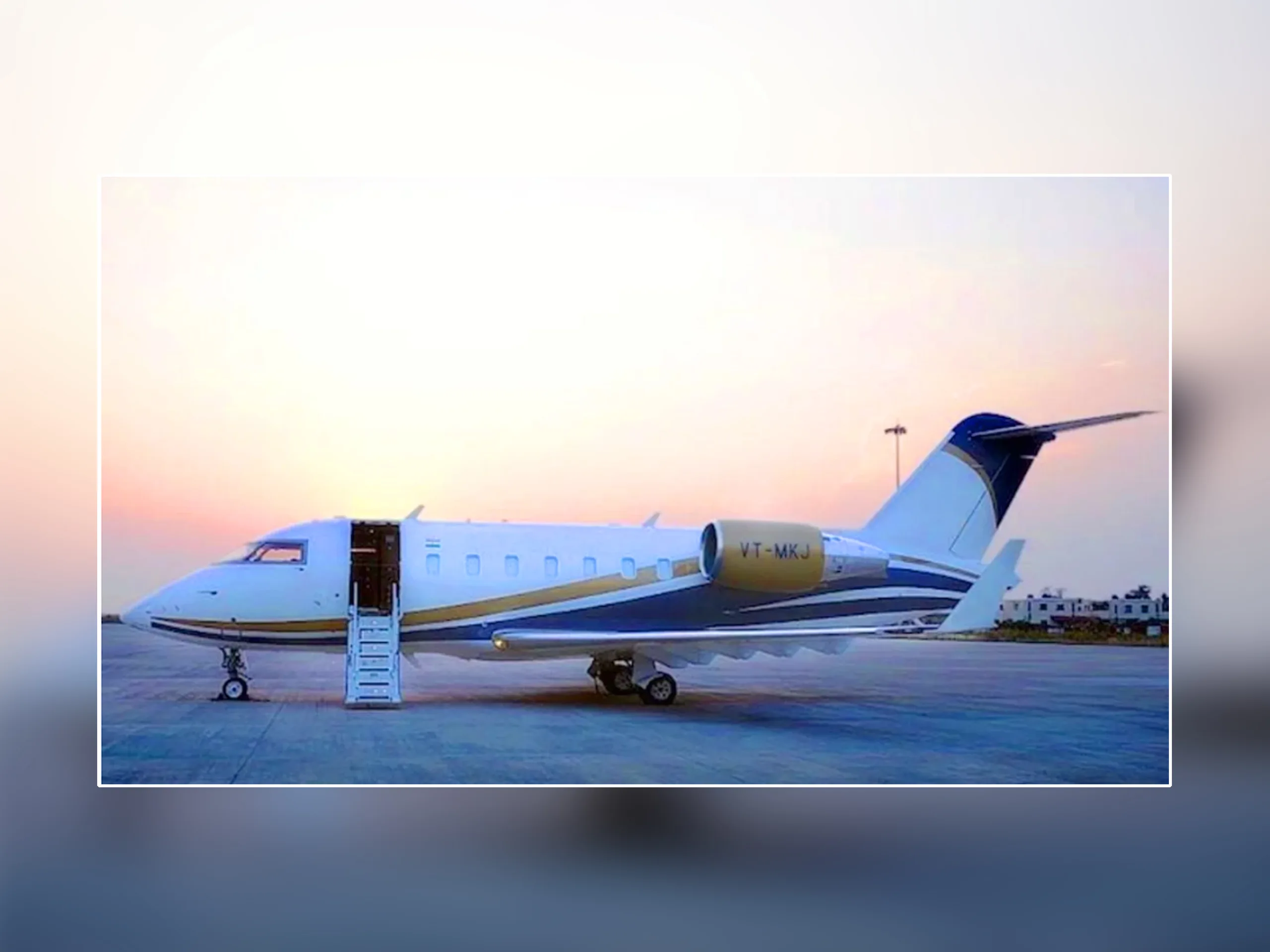 A white and blue Bombardier Challenger business jet with registration VT-MKJ on the tarmac at sunset, with its door open and stairs extended.