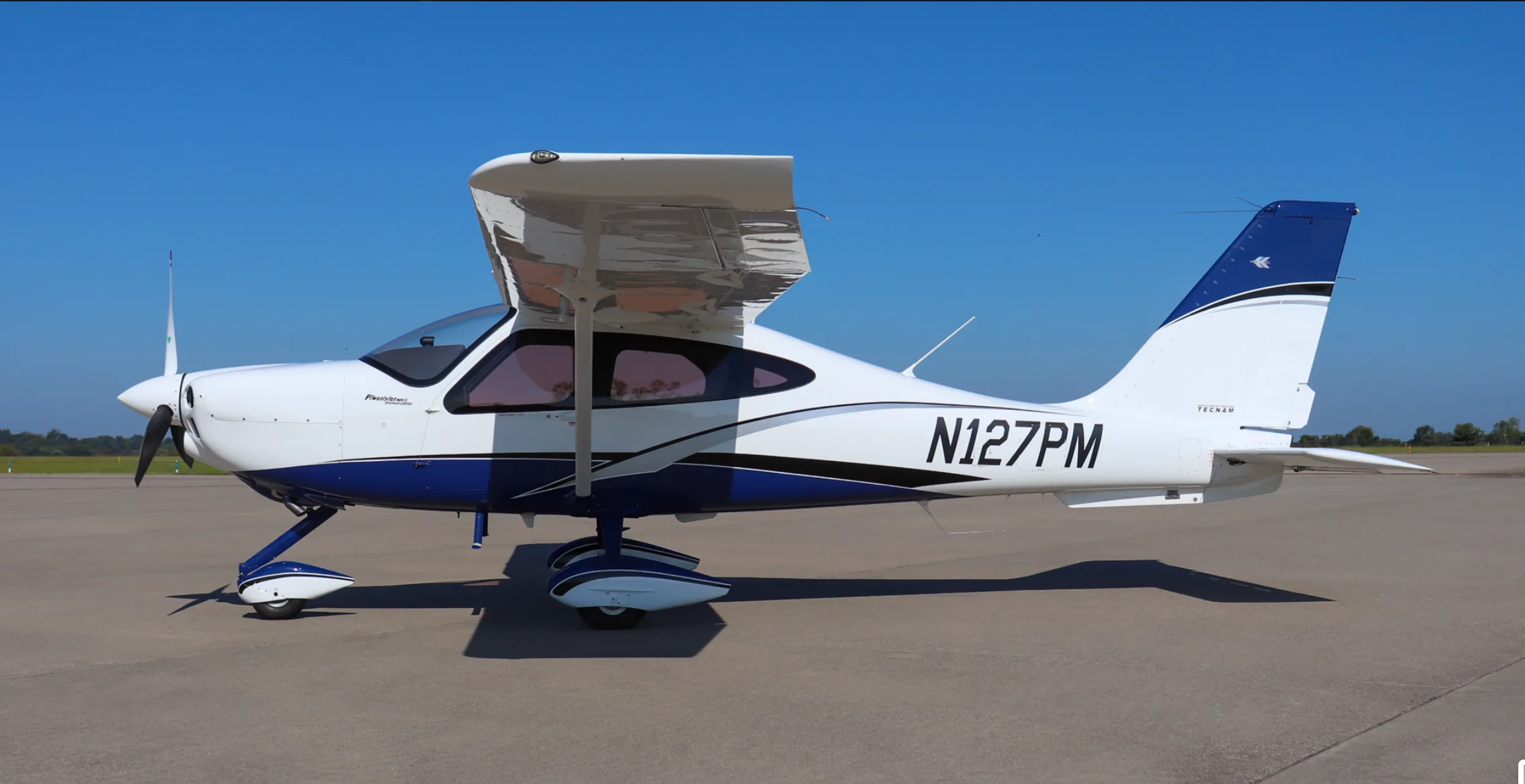 A side view of the white and blue Tecnam P2010 aircraft, registration N127PM, with its doors open, on an airfield.