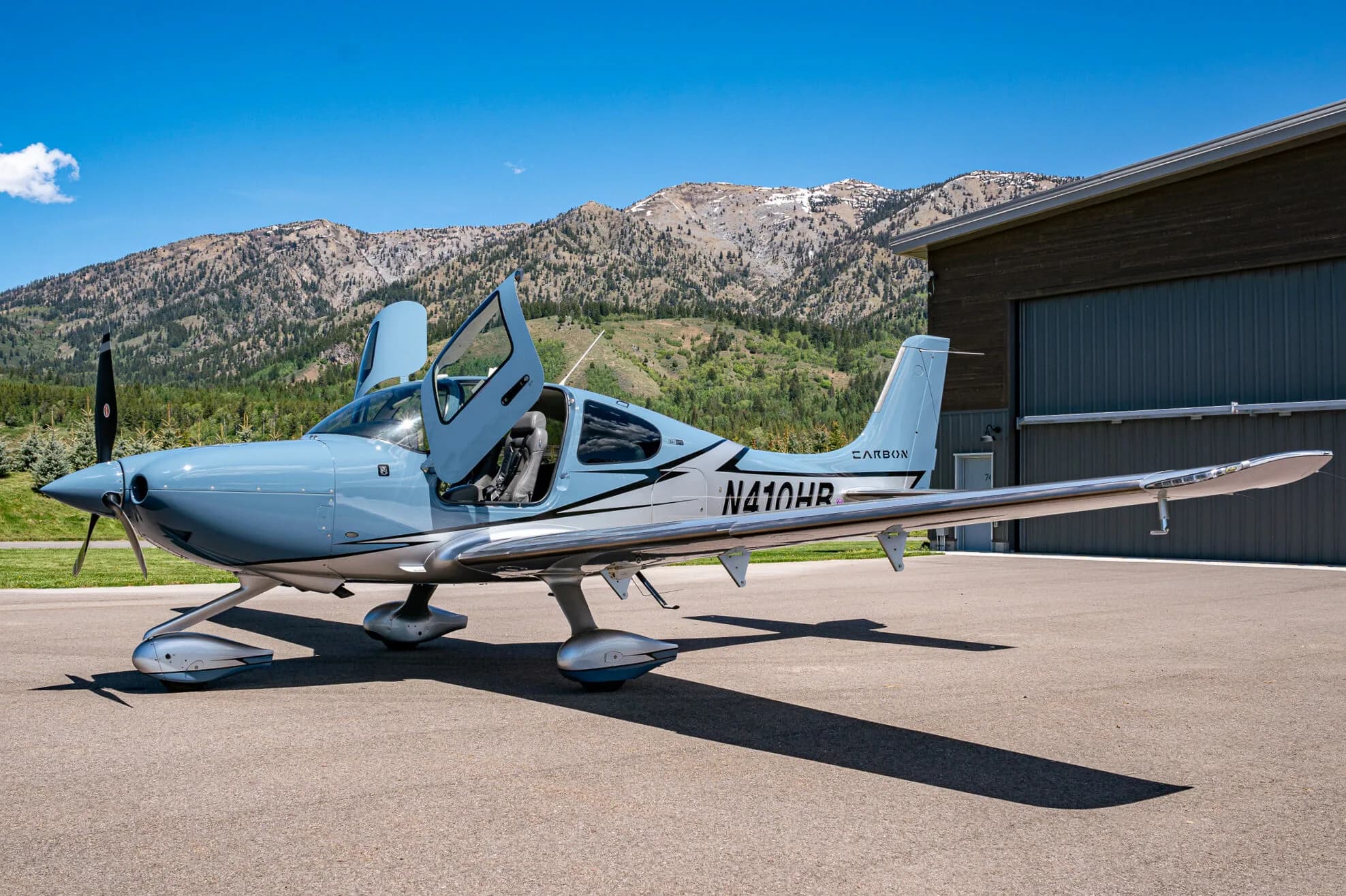 A light blue and silver Cirrus SR22T aircraft (N410HB) with its doors open, parked next to a hangar.