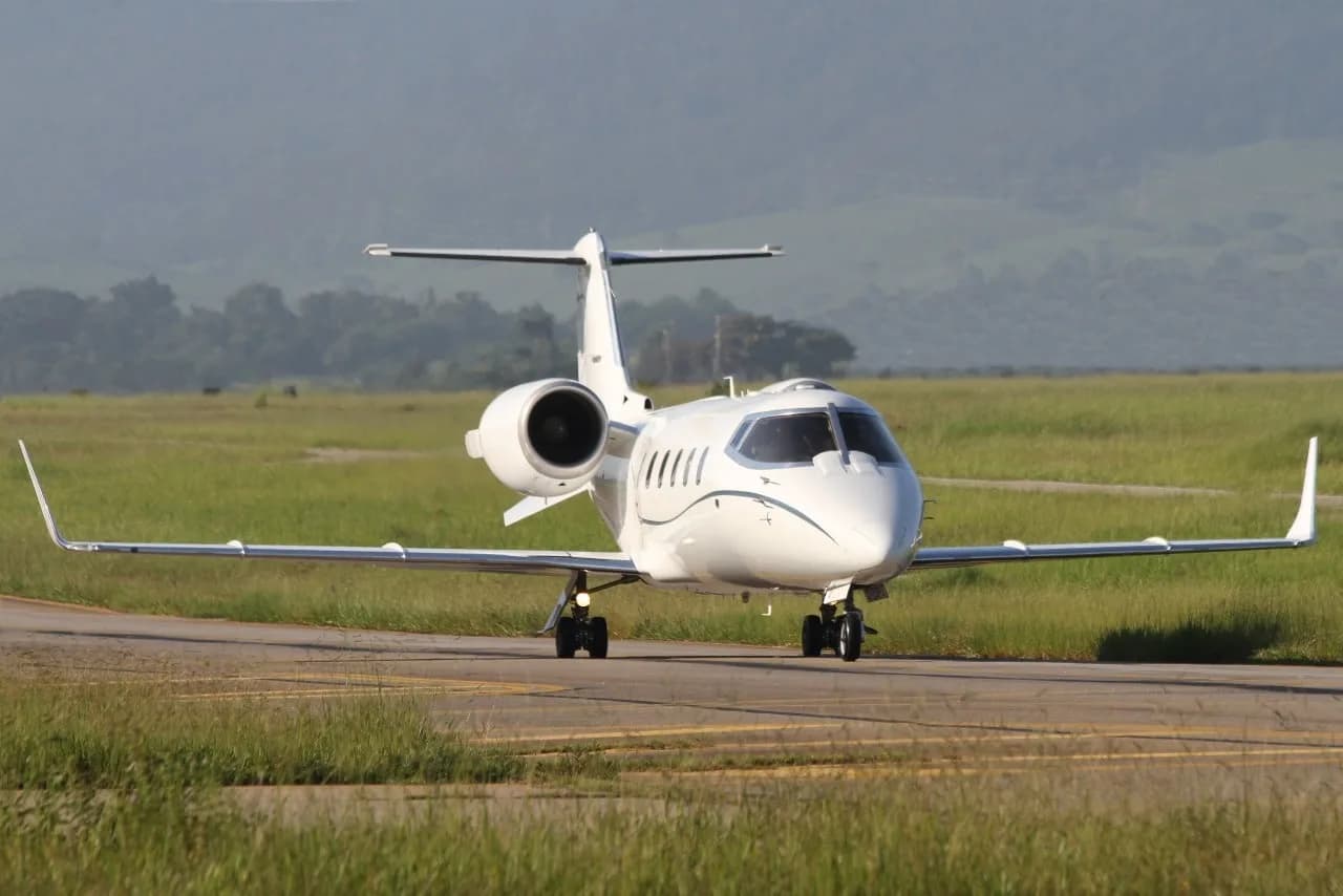 A white business jet with blue pinstripes taxis on a runway with green grass and hills in the background.