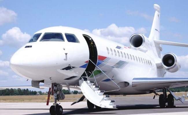 A white Dassault Falcon business jet with a colorful tail design and open passenger door with stairs extended is parked on an airfield.