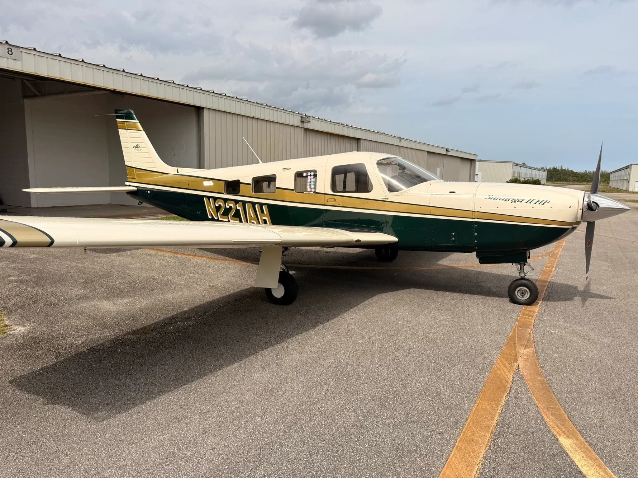Side view of a Piper Saratoga II HP, N221AH, with a white, gold, and dark green paint scheme, parked on an airport tarmac.