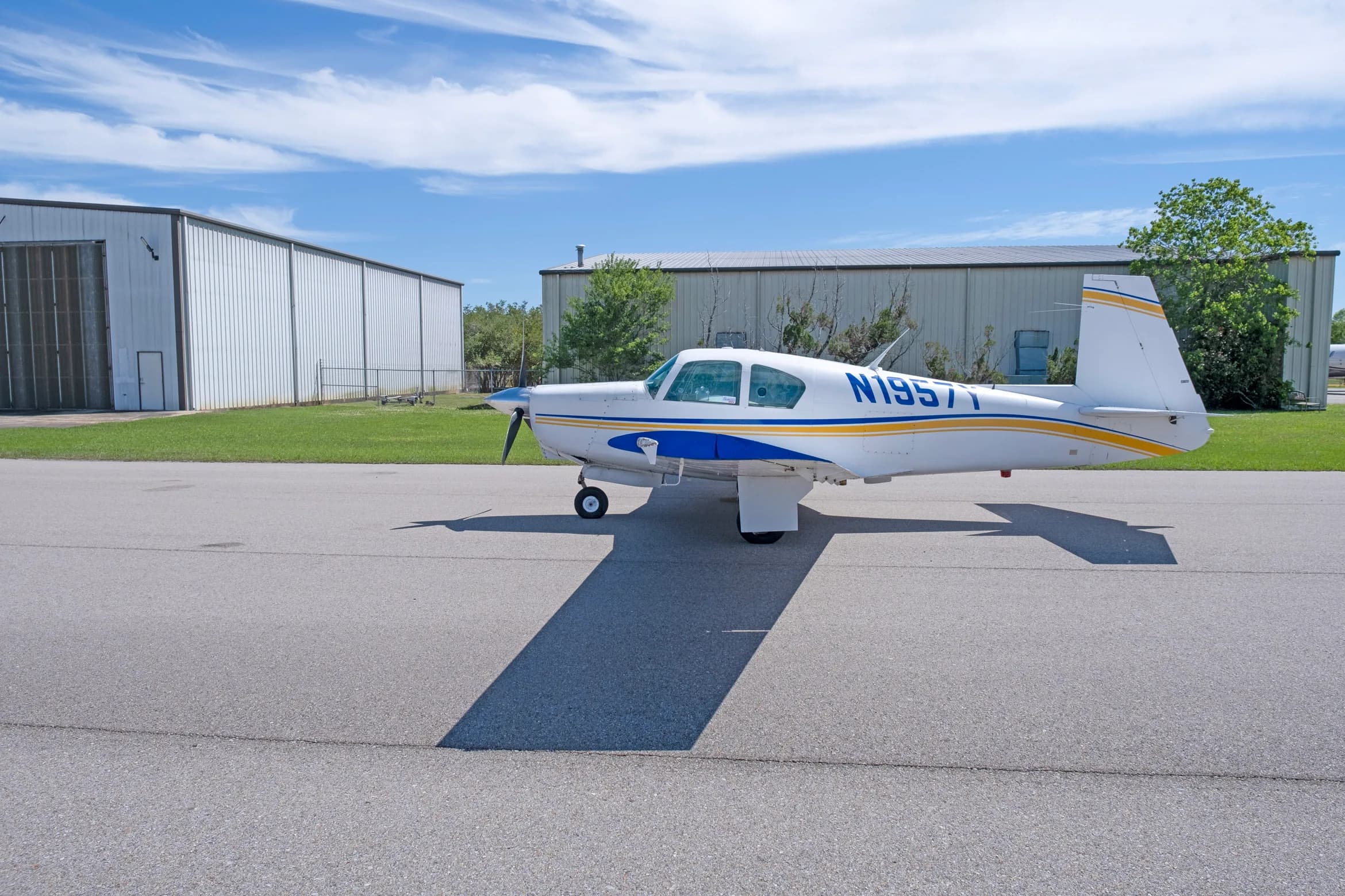 A side view of a white single-engine propeller aircraft with blue and yellow stripes, registration N1957Y, parked on an asphalt apron.