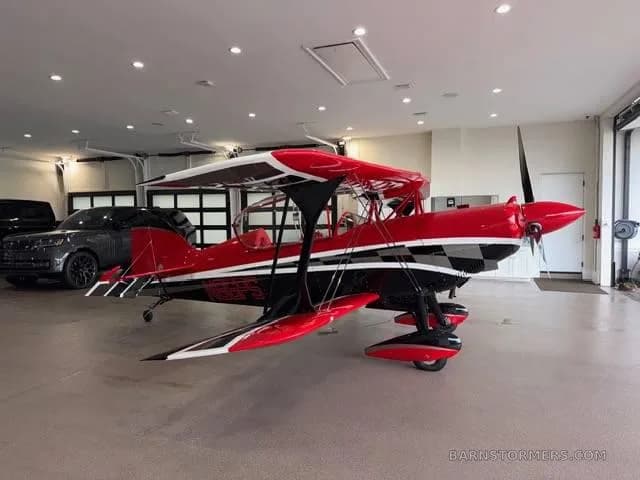 A red and black biplane with a checkered pattern and registration "N86PS" on the fuselage, parked in a garage.