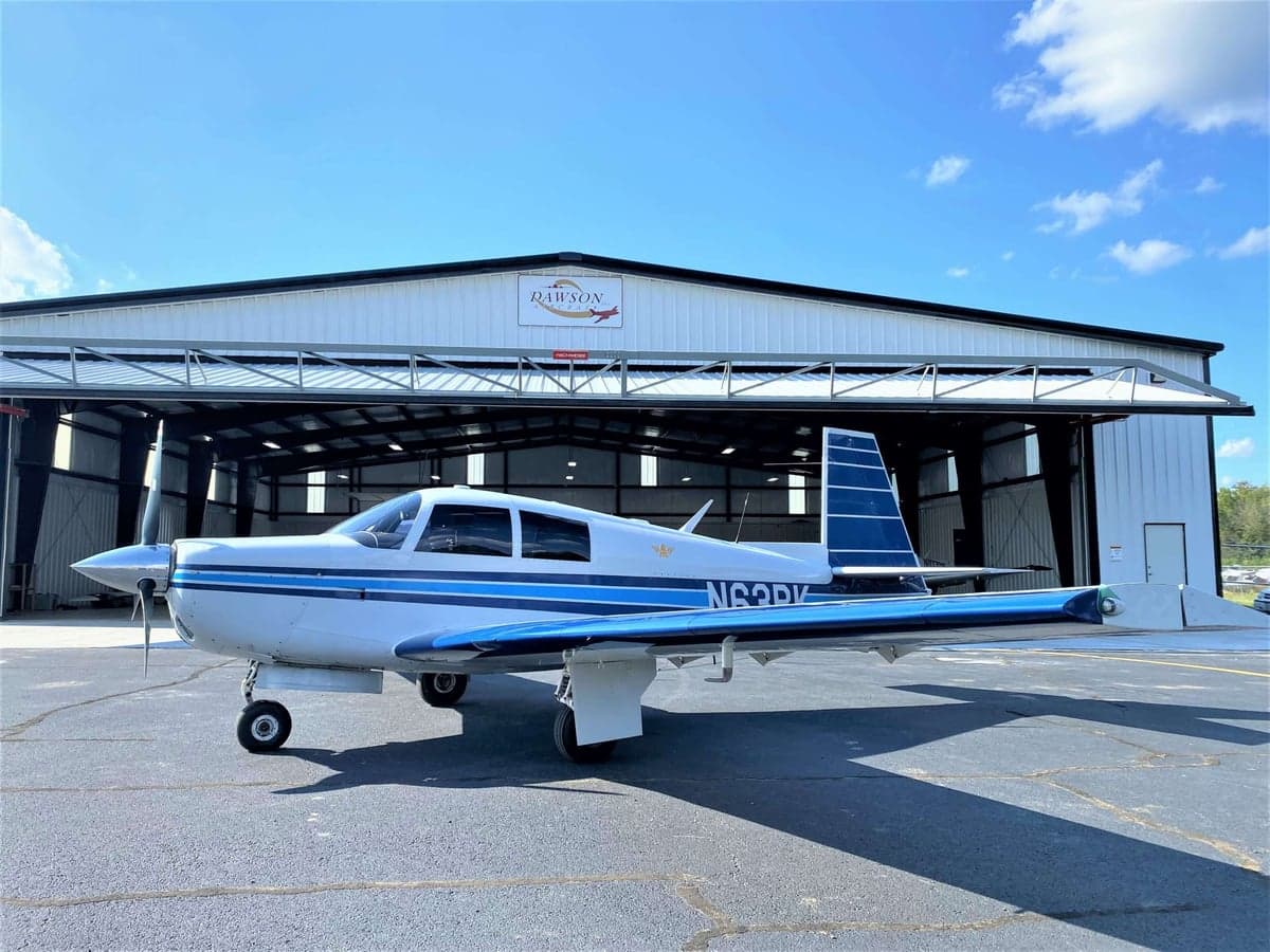 A white and blue Mooney M20C aircraft, tail number N63PK, parked in front of an open hangar on a sunny day.