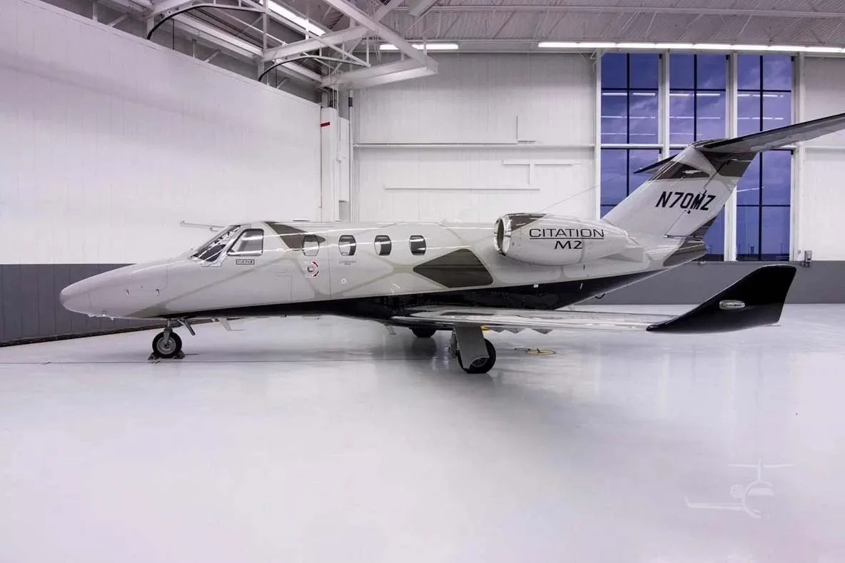 A side view of a silver and black Cessna Citation M2 jet (N70MZ) parked inside a brightly lit hangar.