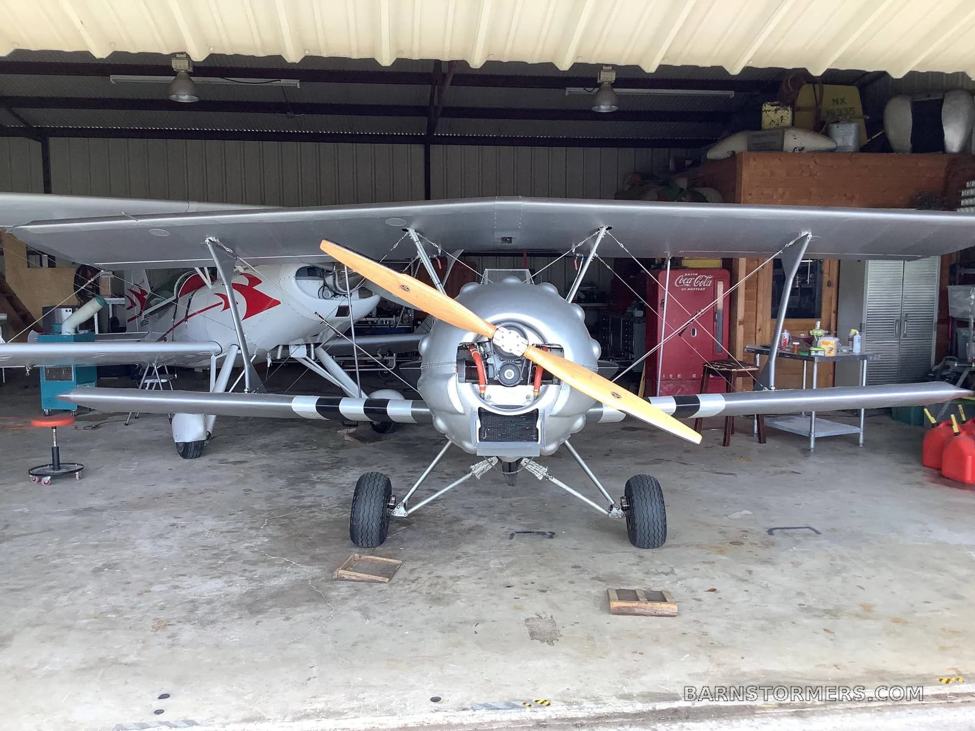 A silver biplane with a wooden propeller is parked head-on in a hangar, with another aircraft behind it.