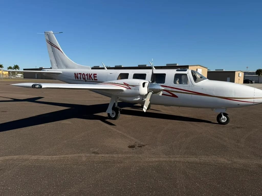 A white twin-engine propeller aircraft with red and gold stripes, registration N701KE, parked on an asphalt ramp with hangars in the background.