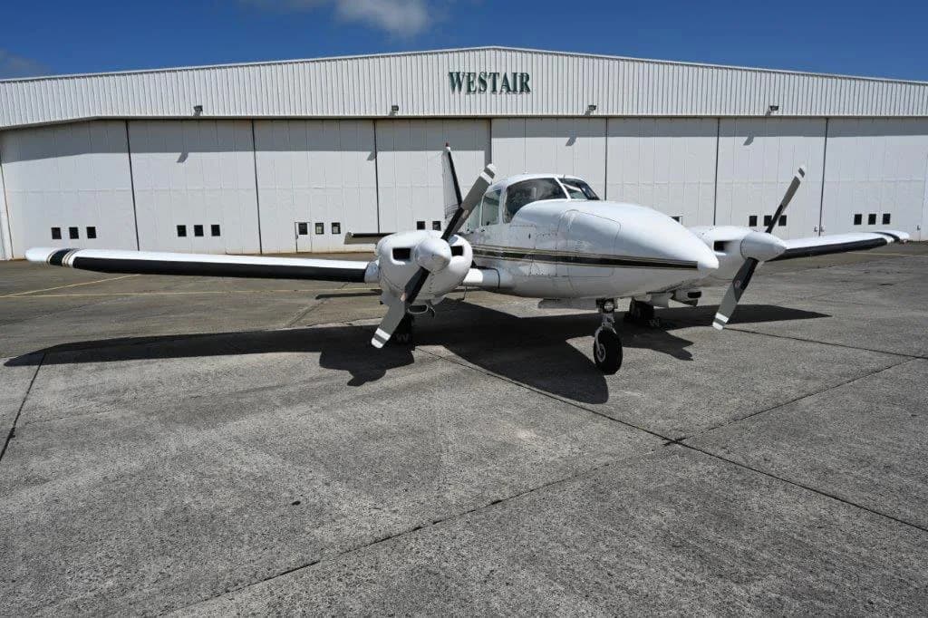 A white twin-engine propeller aircraft with black propellers, viewed from a low angle front-right, parked on a concrete apron in front of a white "WESTAIR" hangar.