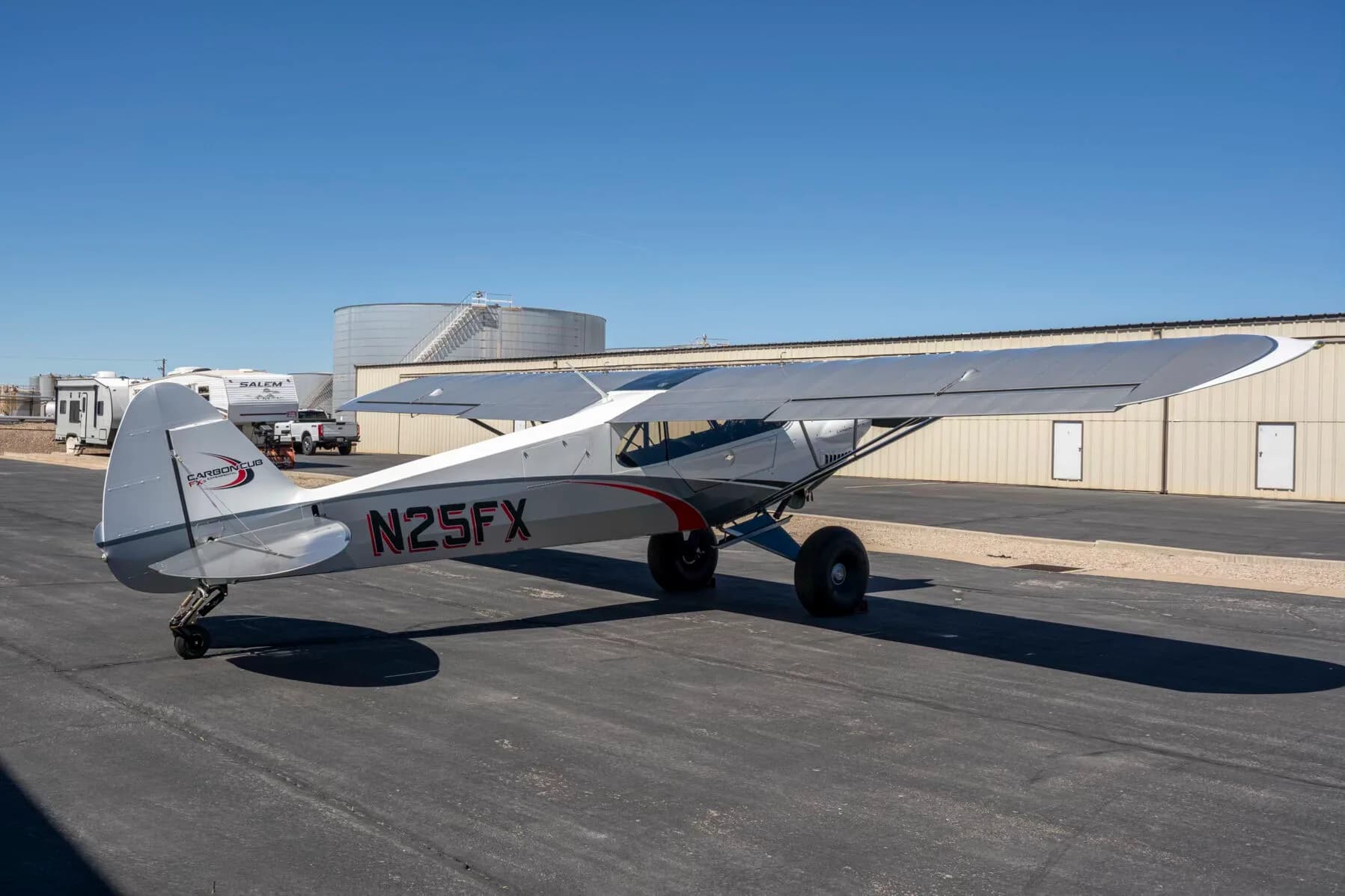 A three-quarter rear view of the white and grey Carbon Cub FX-3 aircraft N25FX parked on an asphalt ramp with storage tanks and a trailer in the background.