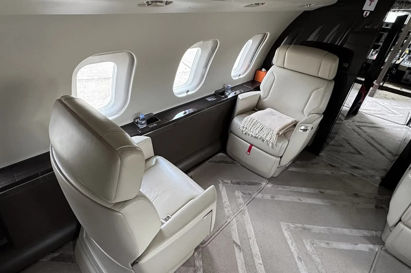 A view inside a private jet cabin, showing two light-colored leather seats, one with a blanket, next to large windows.