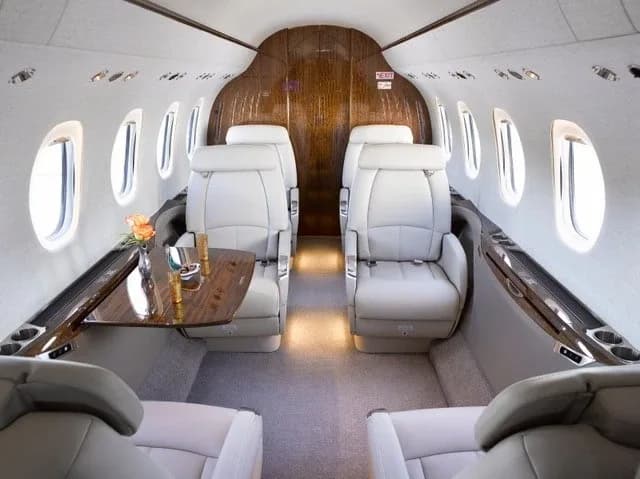 The luxurious interior cabin of a business jet with light grey leather seats, a wooden table, and large windows.