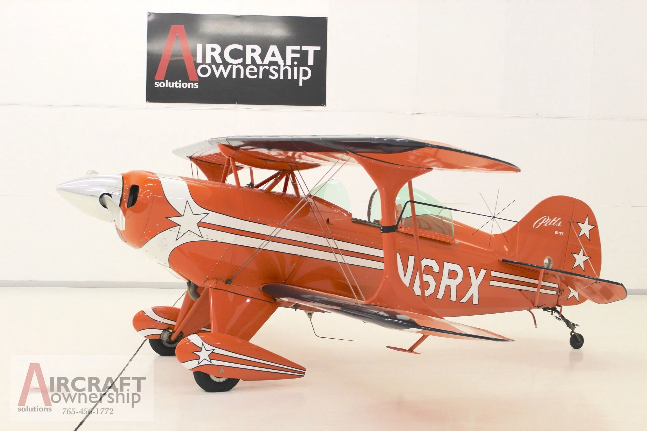 A side view of an orange Pitts S-1T biplane with white stripes and stars, parked in a hangar.