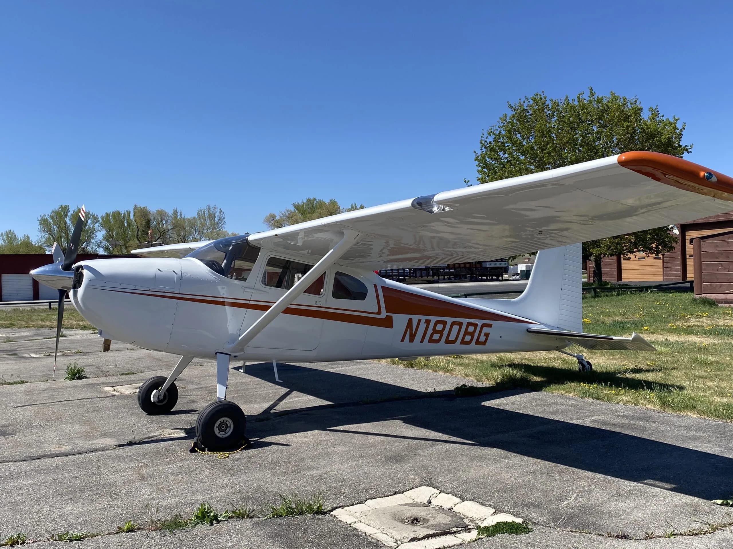 A white Cessna 170B with orange stripes and N180BG registration parked on an airfield under a clear blue sky.