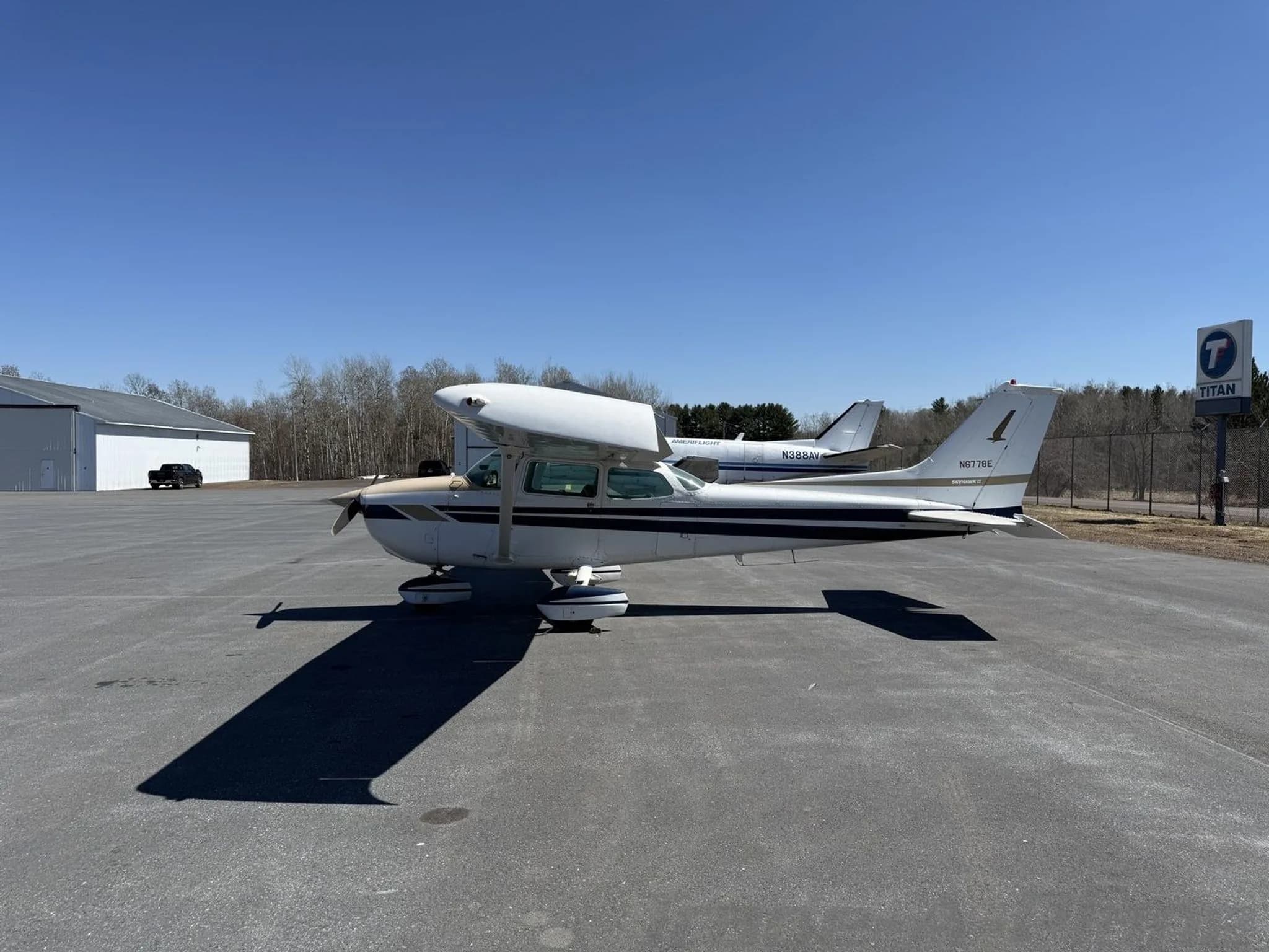 A white and tan Cessna 172 aircraft N6778E with blue stripes parked on an asphalt apron with another aircraft in the background.