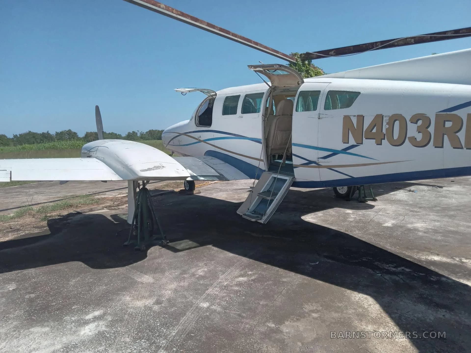 A white and blue twin-engine propeller aircraft with registration N403RM, parked on an asphalt surface with its passenger door open, under a clear blue sky.