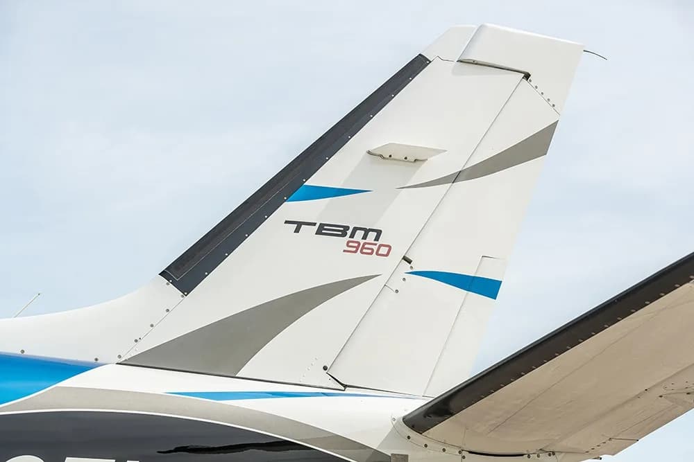A close-up of the TBM 960 aircraft's vertical stabilizer, showing the TBM 960 logo and blue and gray accents.