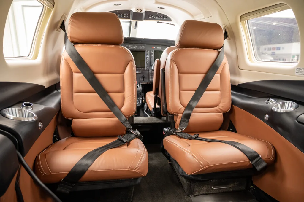 The aircraft cabin interior, showing two tan leather passenger seats with seatbelts and a glimpse of the cockpit beyond.