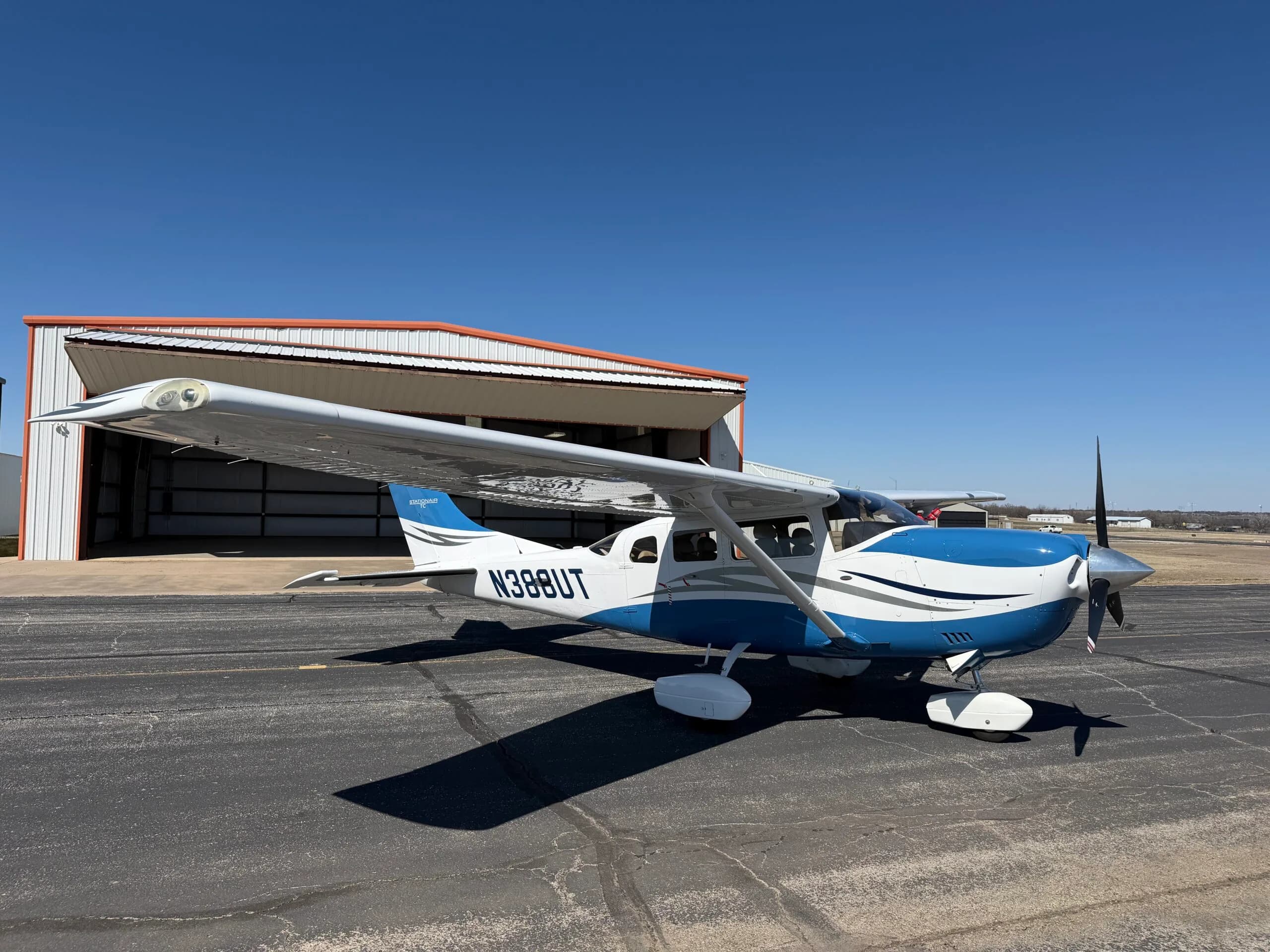 A white and blue single-engine propeller aircraft with registration N388UT is parked on an asphalt ramp in front of an open hangar under a clear blue sky.