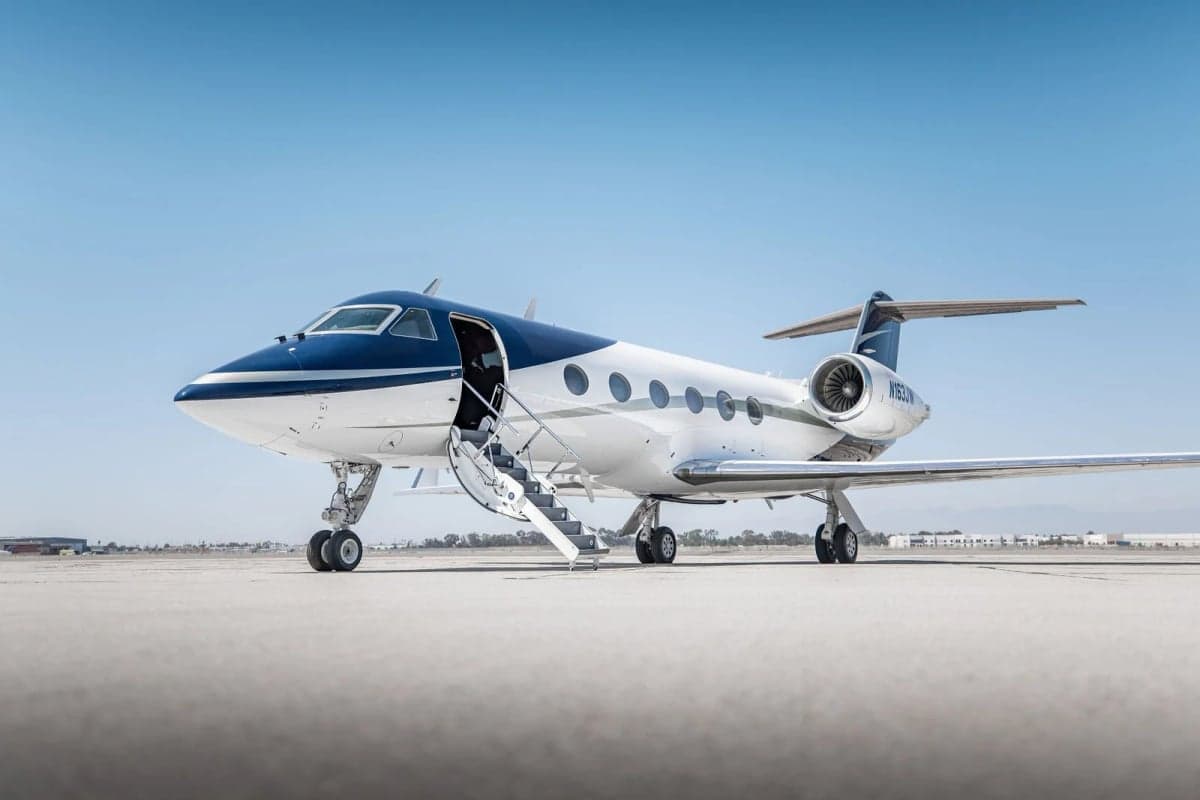 A white and blue Gulfstream G550 business jet with its door open and stairs extended, parked on a sunny tarmac.