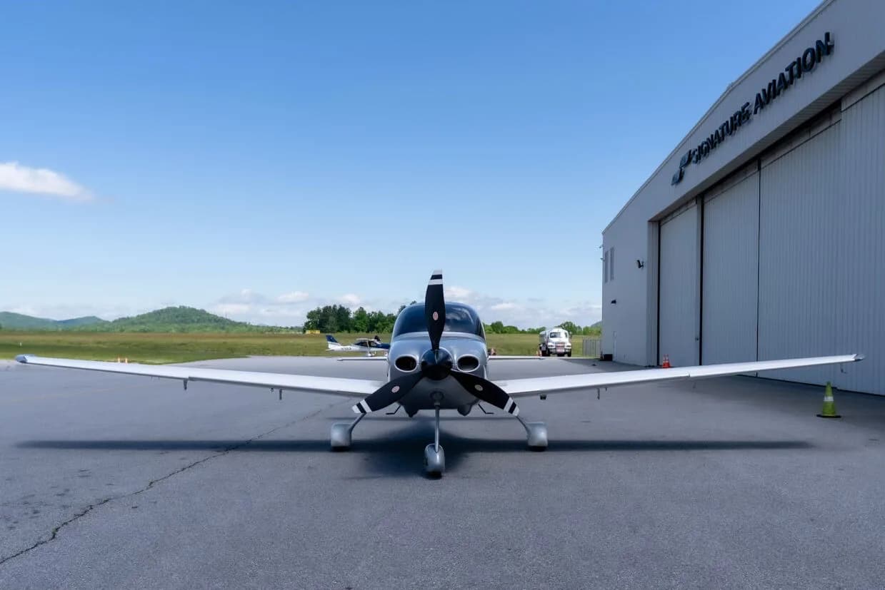 A head-on view of a silver and white Cirrus SR22T aircraft parked in front of a "Signature Aviation" hangar.