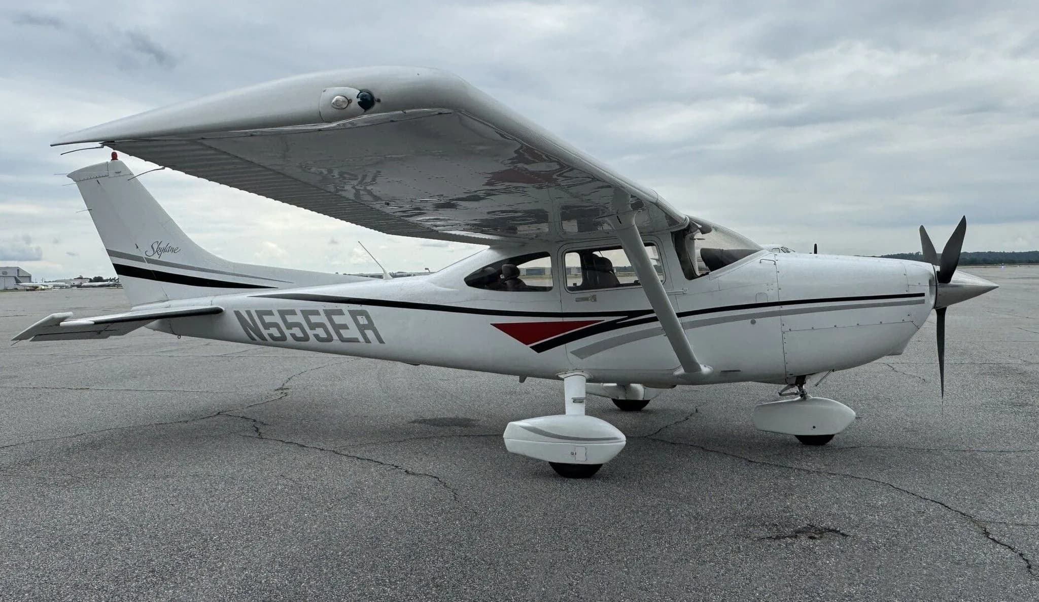 A white Cessna 182 Skylane with black, grey, and red stripes parked on an airfield under a cloudy sky.