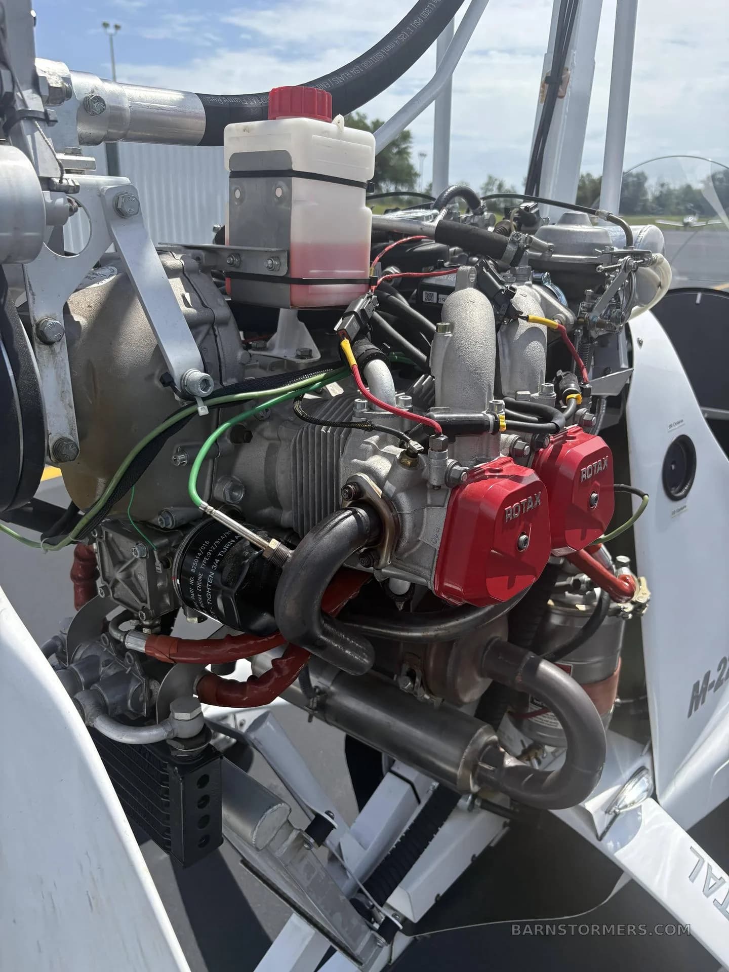 A detailed view of the exposed Rotax engine of the M-22 gyrocopter, featuring red valve covers and various hoses.