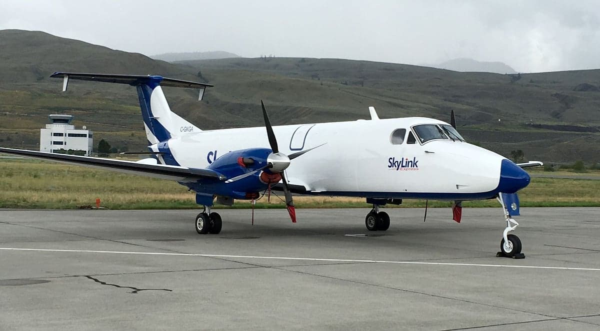 A white and blue SkyLink turboprop aircraft (Beechcraft 1900D) parked on an airport tarmac with a control tower and hills in the background.