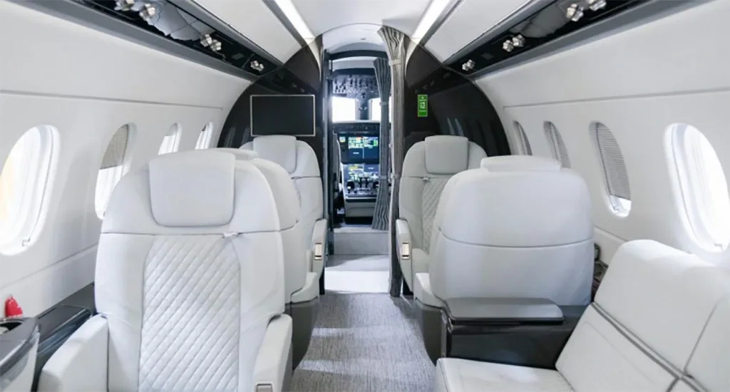 The elegant interior of a business jet cabin with light grey seats and a view into the cockpit.