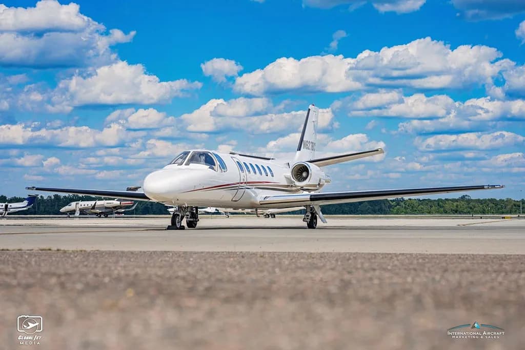 A low-angle front-side view of a white Cessna Citation Bravo business jet with red and black stripes on an airport tarmac.