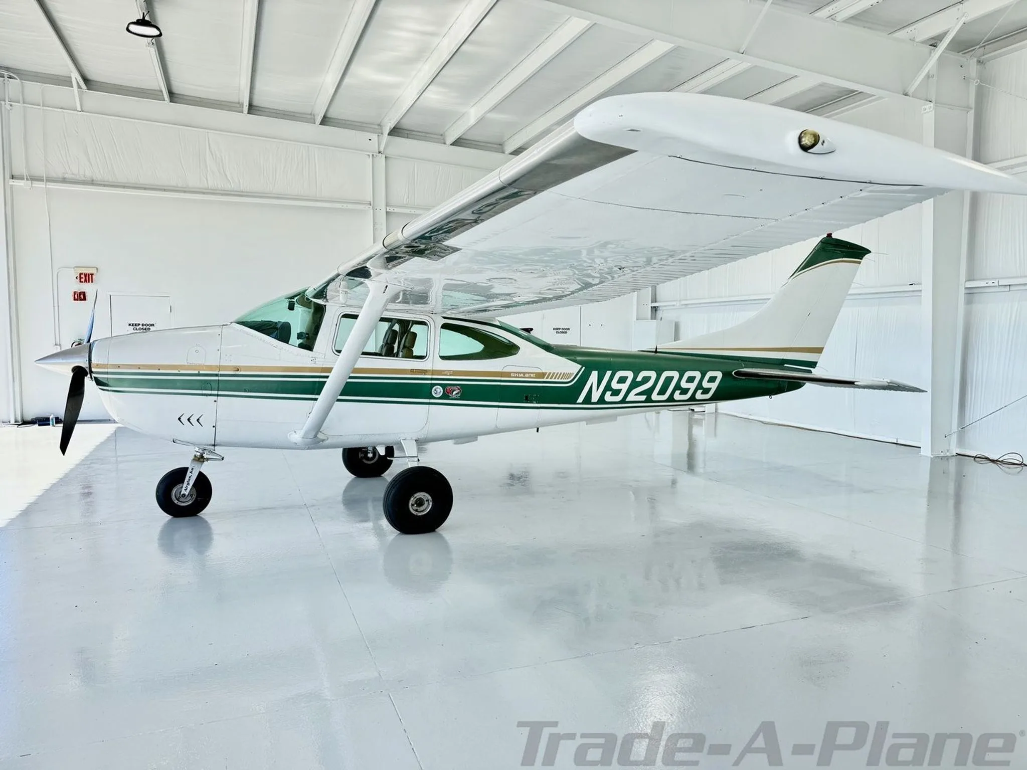 A white and green Cessna Skylane N92099 parked in a brightly lit hangar, viewed from the front-left.