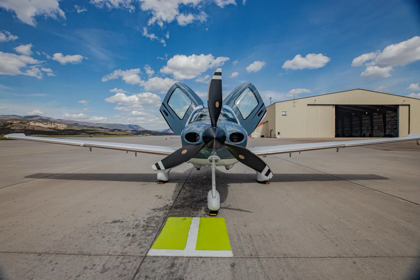 Front view of a blue and white Cirrus SR22T GTS aircraft with its gull-wing doors open, parked on an airport tarmac.