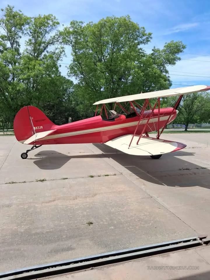 Side view of a red biplane with white stripes and tail number N57JS, parked on a concrete apron with trees in the background.
