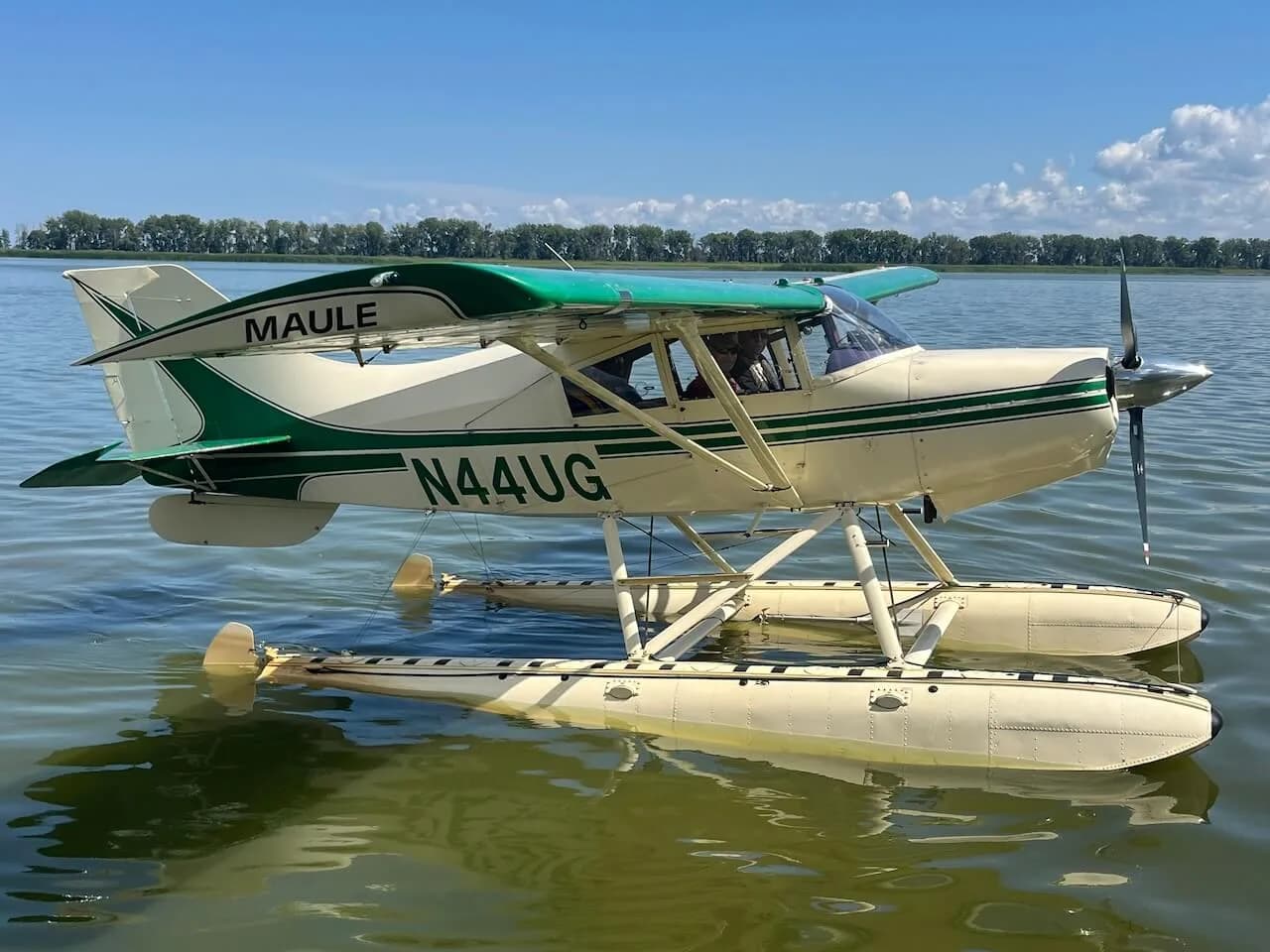 A Maule seaplane with a green and white paint scheme on floats is floating on a lake under a blue sky.