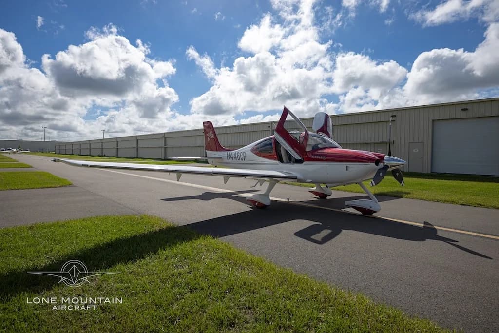 A red and white Cirrus SR22 aircraft with its doors open, parked on an asphalt ramp with hangars in the background under a cloudy sky.