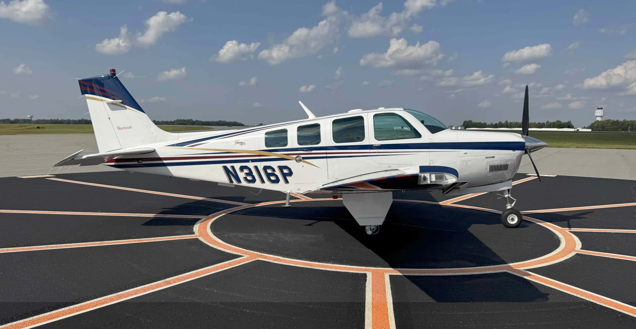 A white Beechcraft Bonanza N316P with blue and gold stripes parked on an airport tarmac.