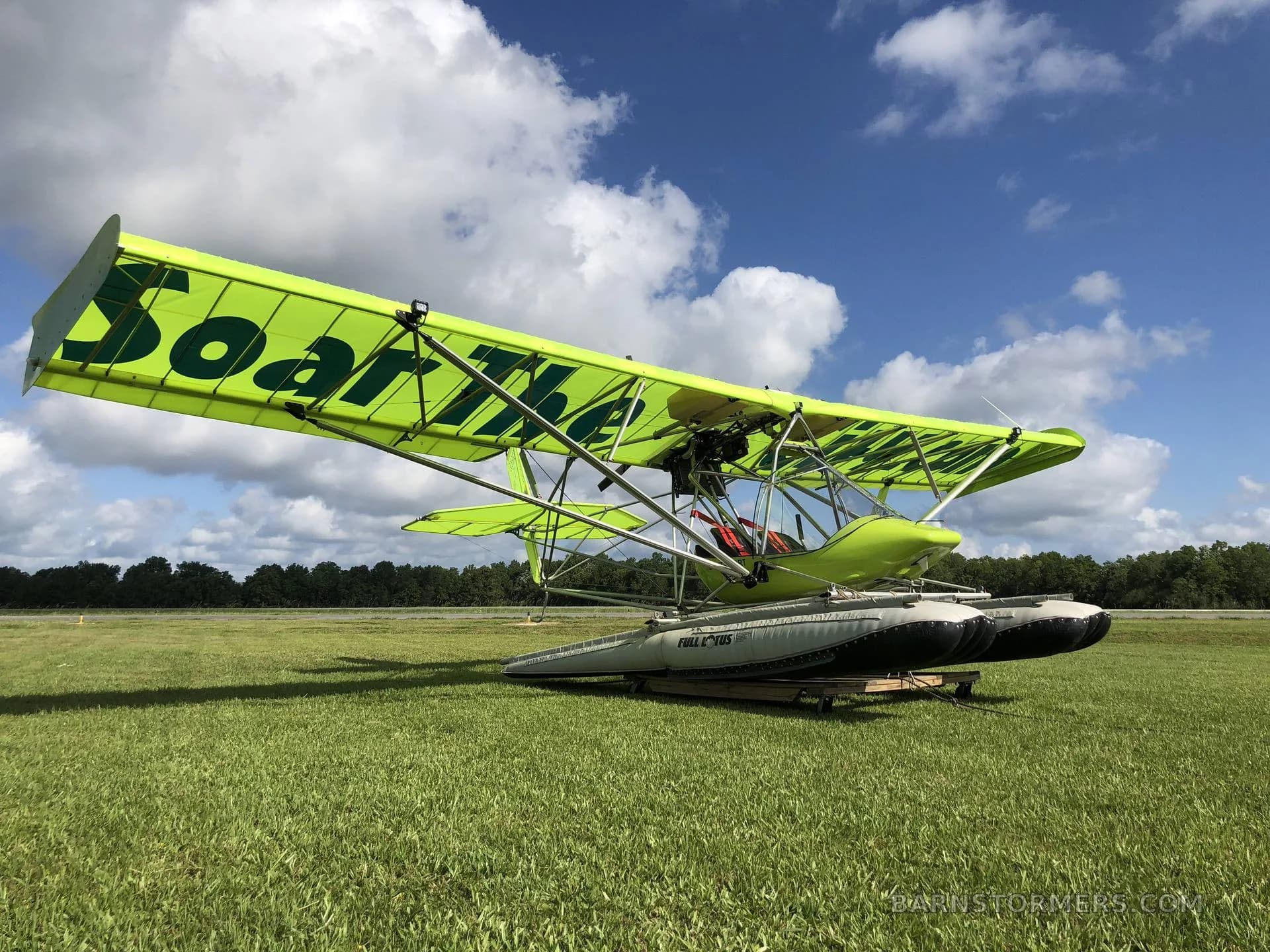 A bright green ultralight seaplane with "Soar The City" on its wings is parked on a grassy airfield under a partly cloudy blue sky.