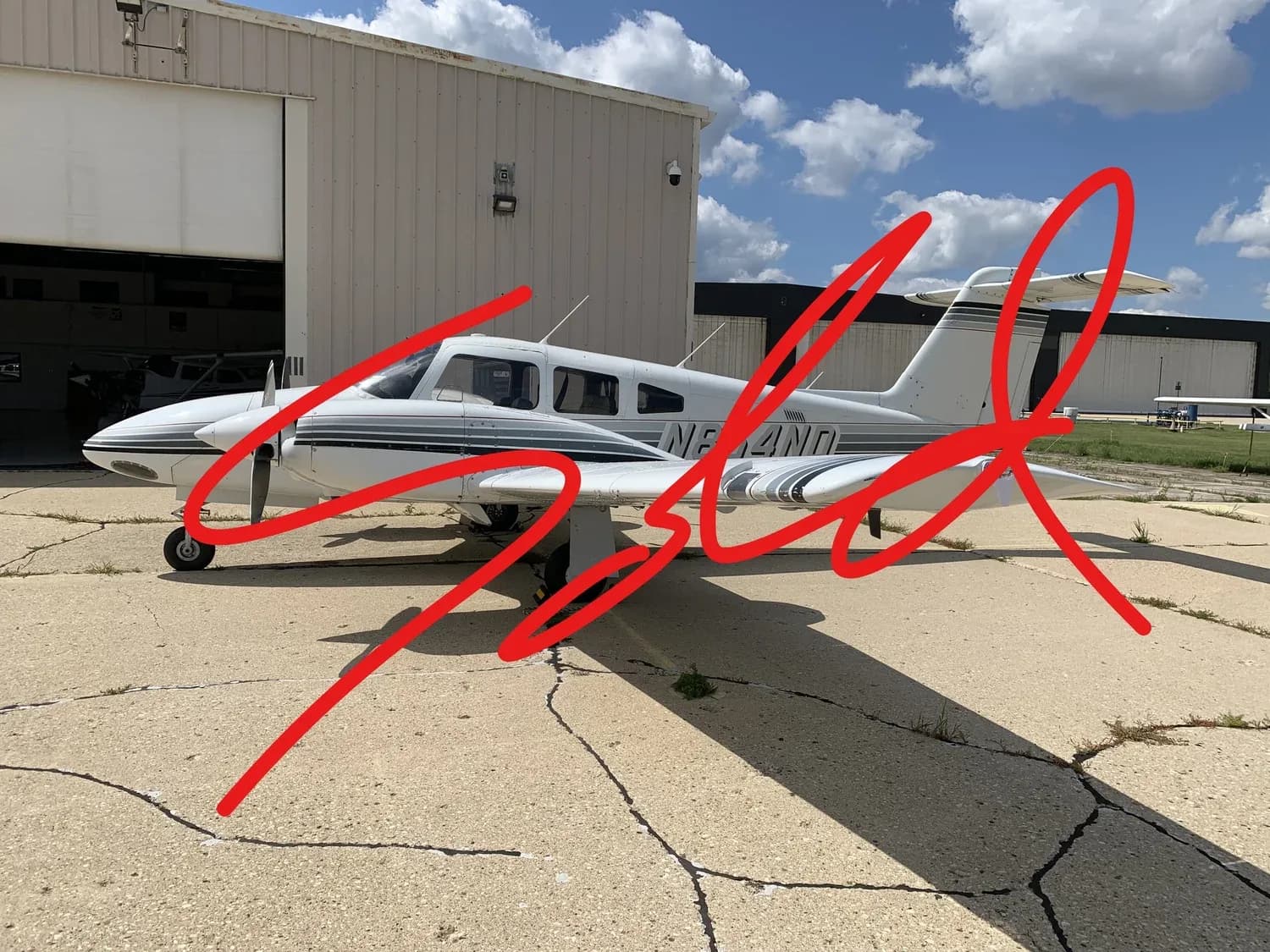 A white Piper Saratoga aircraft with grey stripes parked on a tarmac next to a hangar under a partly cloudy sky.