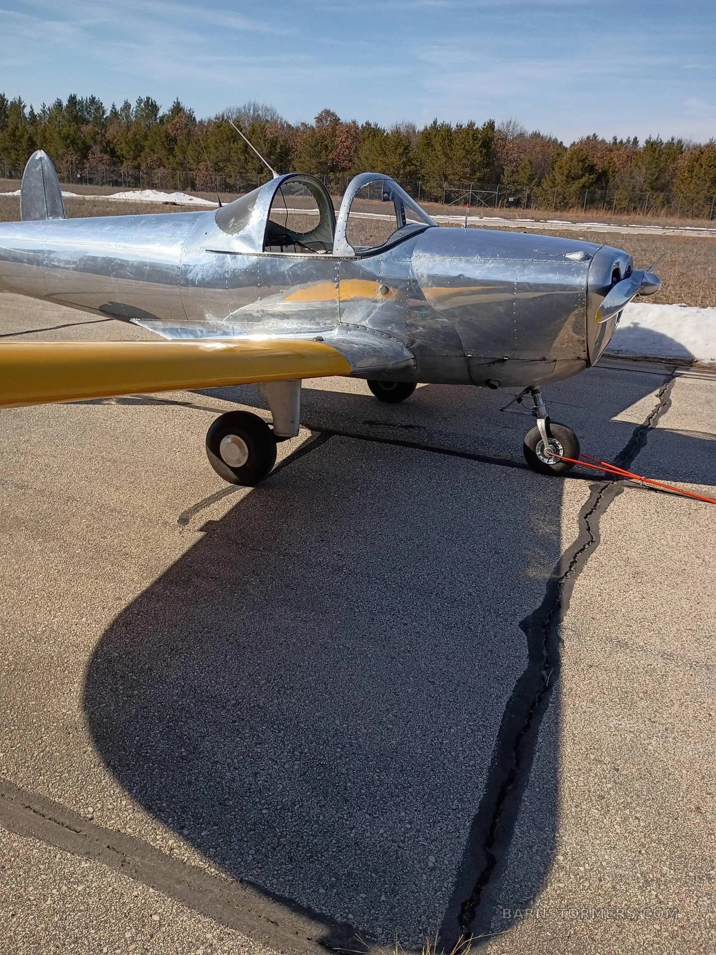 A front-side view of a polished silver aircraft with yellow wings parked on an airfield.