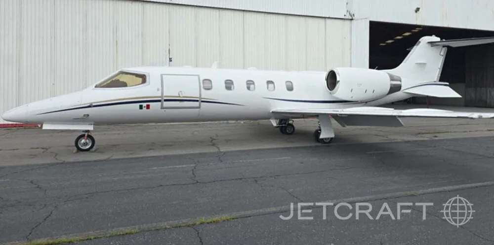 A white business jet with a black and gold stripe and a Mexican flag on the nose, parked in front of a hangar.