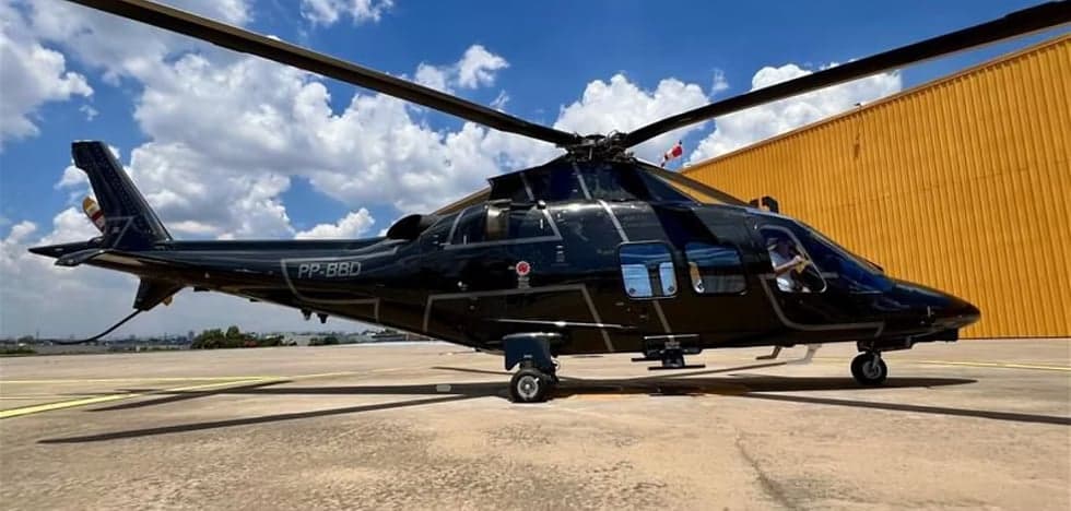 A black helicopter with registration PP-BBD sits on the tarmac in front of a yellow hangar.