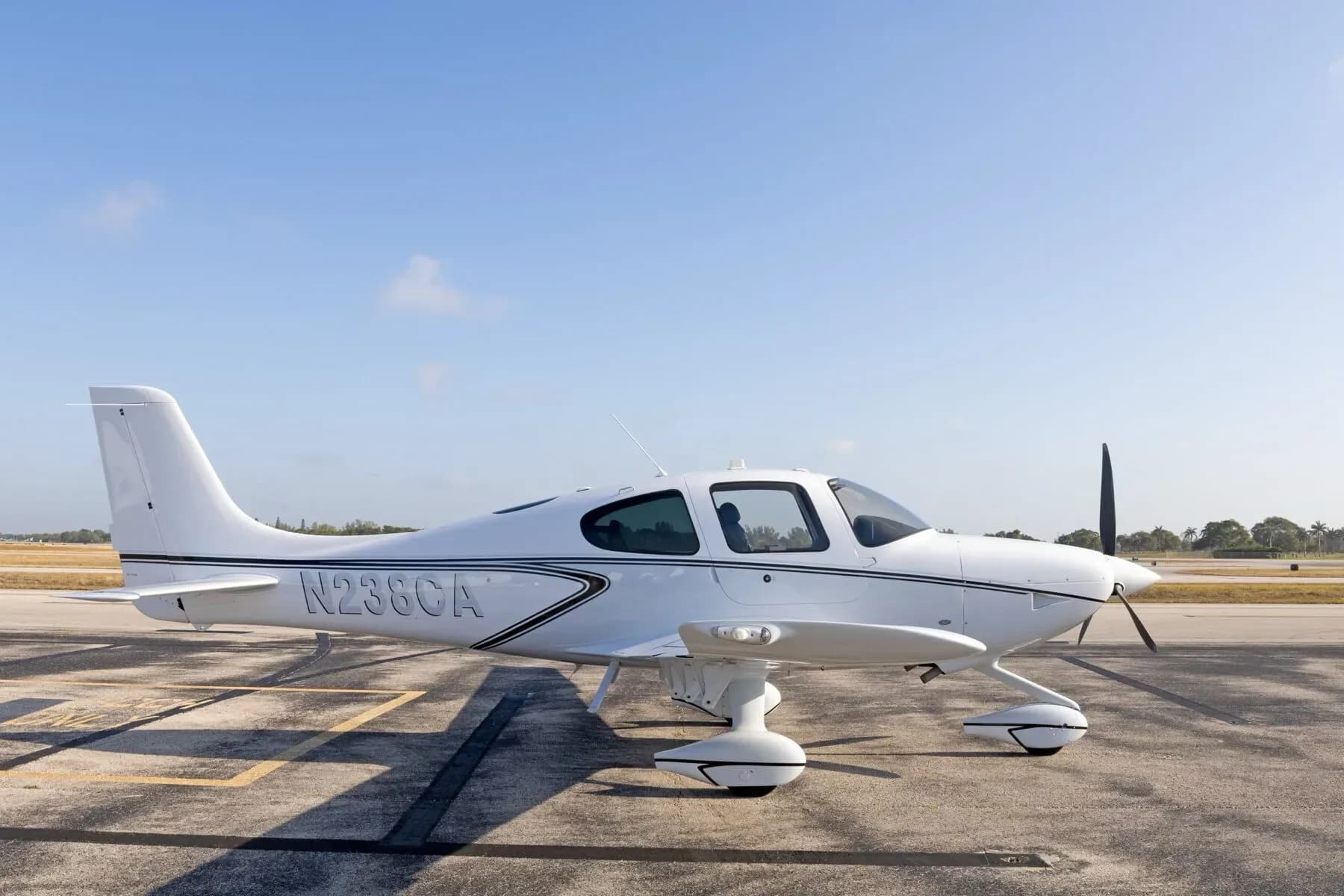 A white Cirrus SR22 aircraft with black stripes is seen from the side, parked on an airport tarmac.