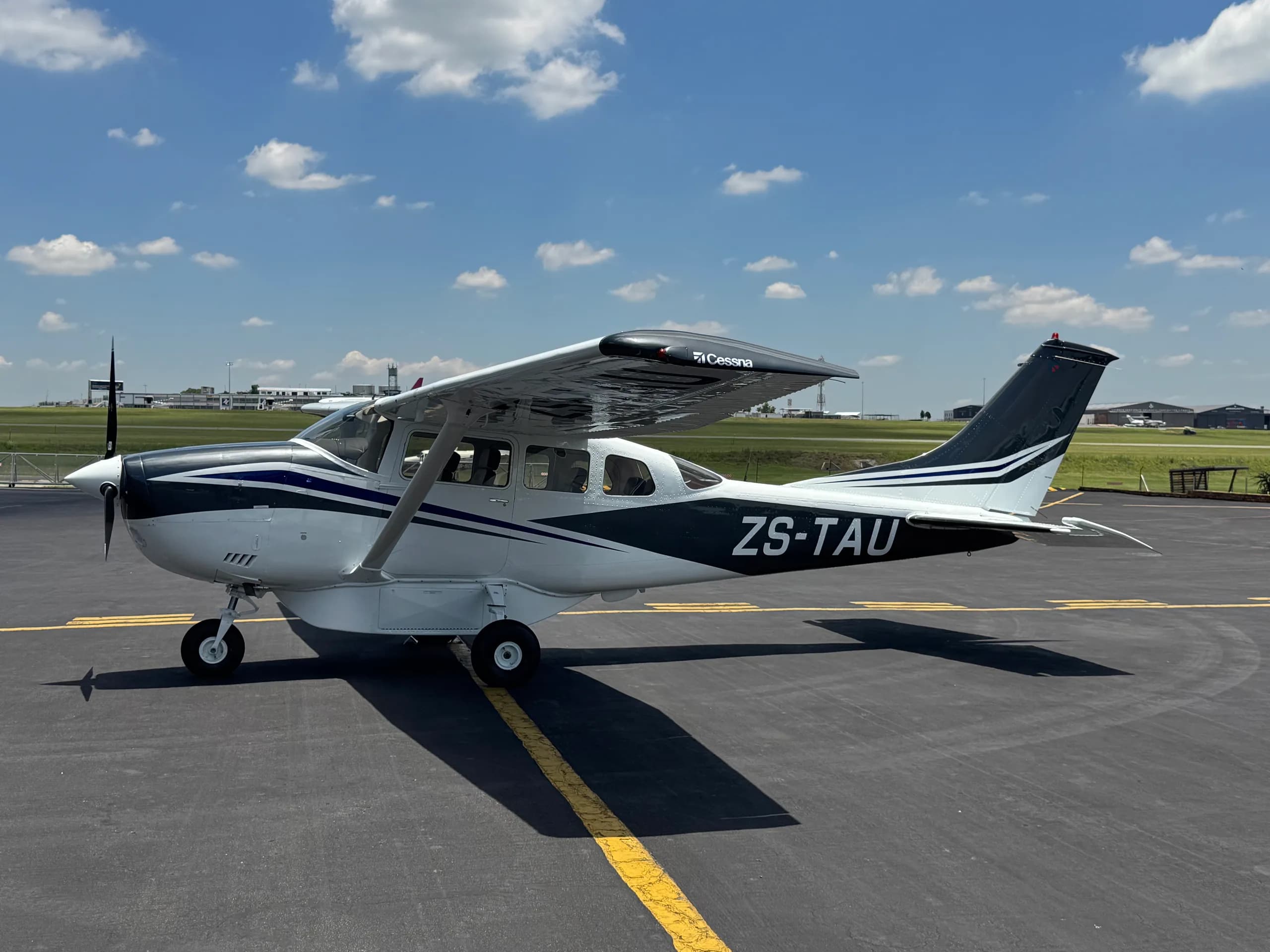 A white and black Cessna 206 parked on an tarmac under a blue sky with clouds.
