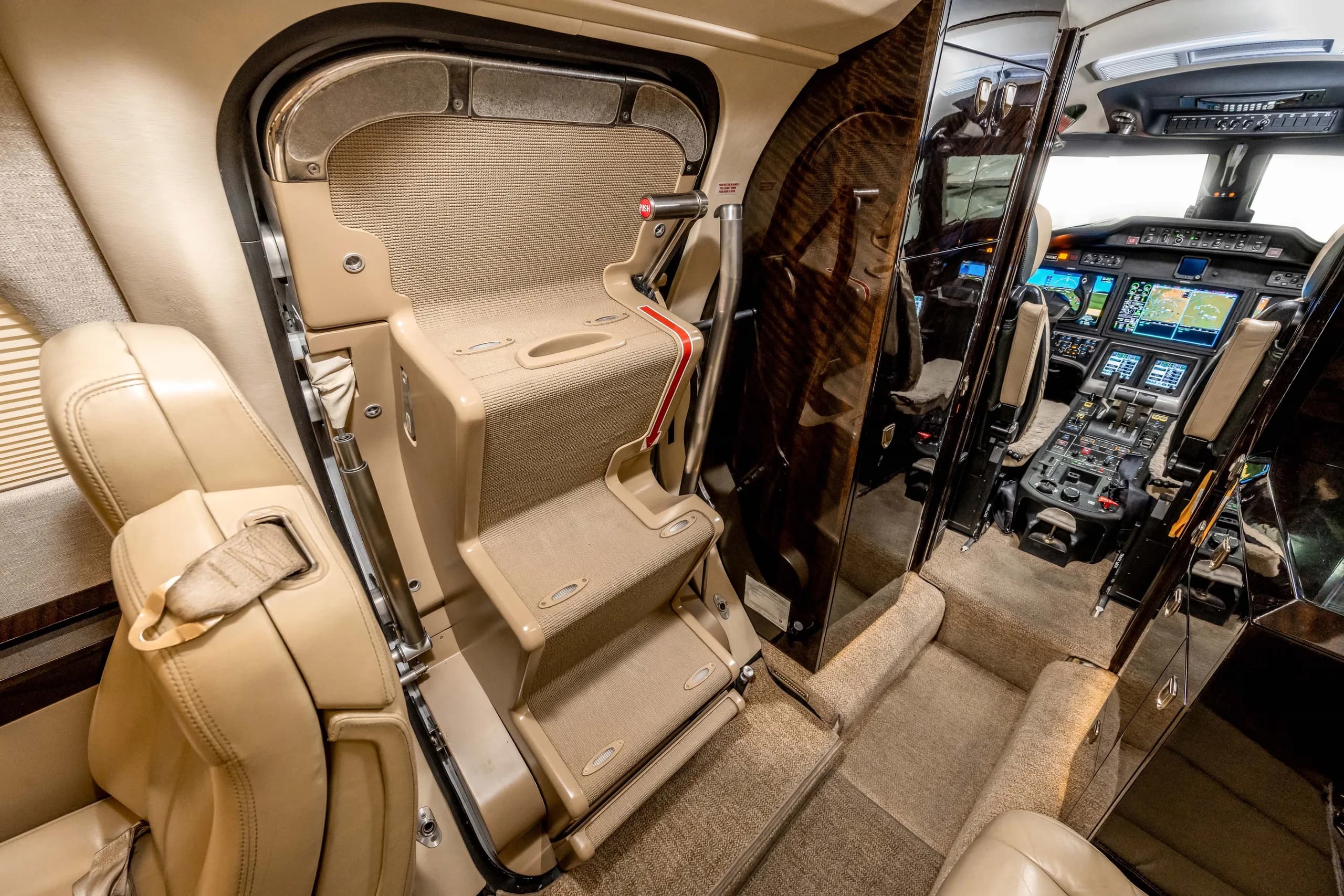 The entry stairs of a business jet, with light tan carpet and dark wood paneling, leading into the cabin and towards the cockpit.